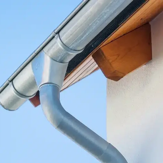Installation de gouttières Gouttière et descente d'eau pluviale en zinc fixées à la toiture. Détail de la corniche en bois et de la façade blanche sous ciel bleu.
